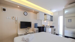 A hotel room with a bed, a TV, a water dispenser, a clock, and a small kitchenette. - Newly Furnished Studio at Bassura City Apartment (Jakarta)