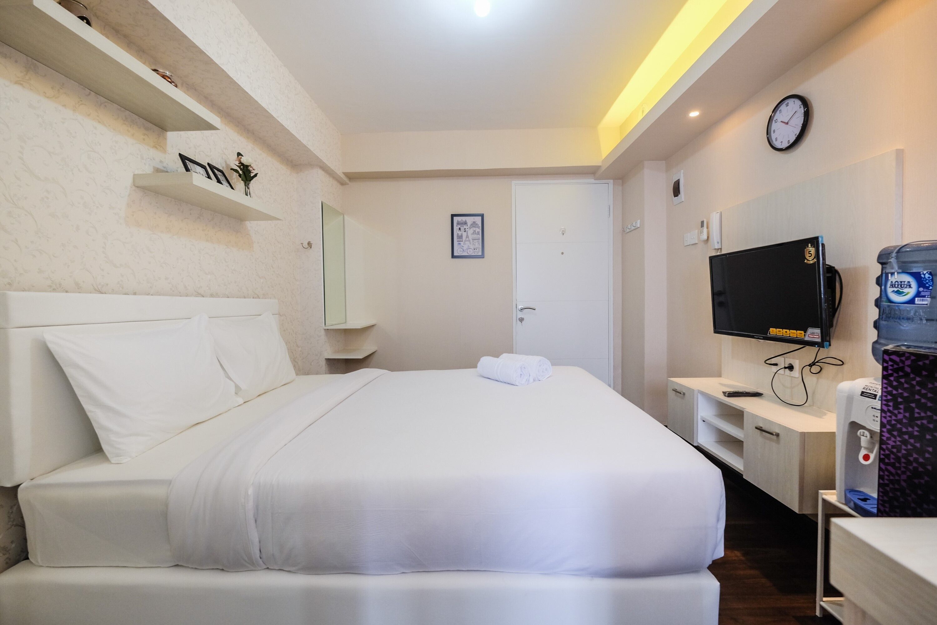 A hotel room with a bed, a TV, a water dispenser, and a clock on the wall.