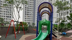 A playground with a green slide, red swings, and a climbing structure, surrounded by a landscaped area with trees and a building in the background. - Comfortable Studio Apartment M-Town Residence (Gading Serpong)