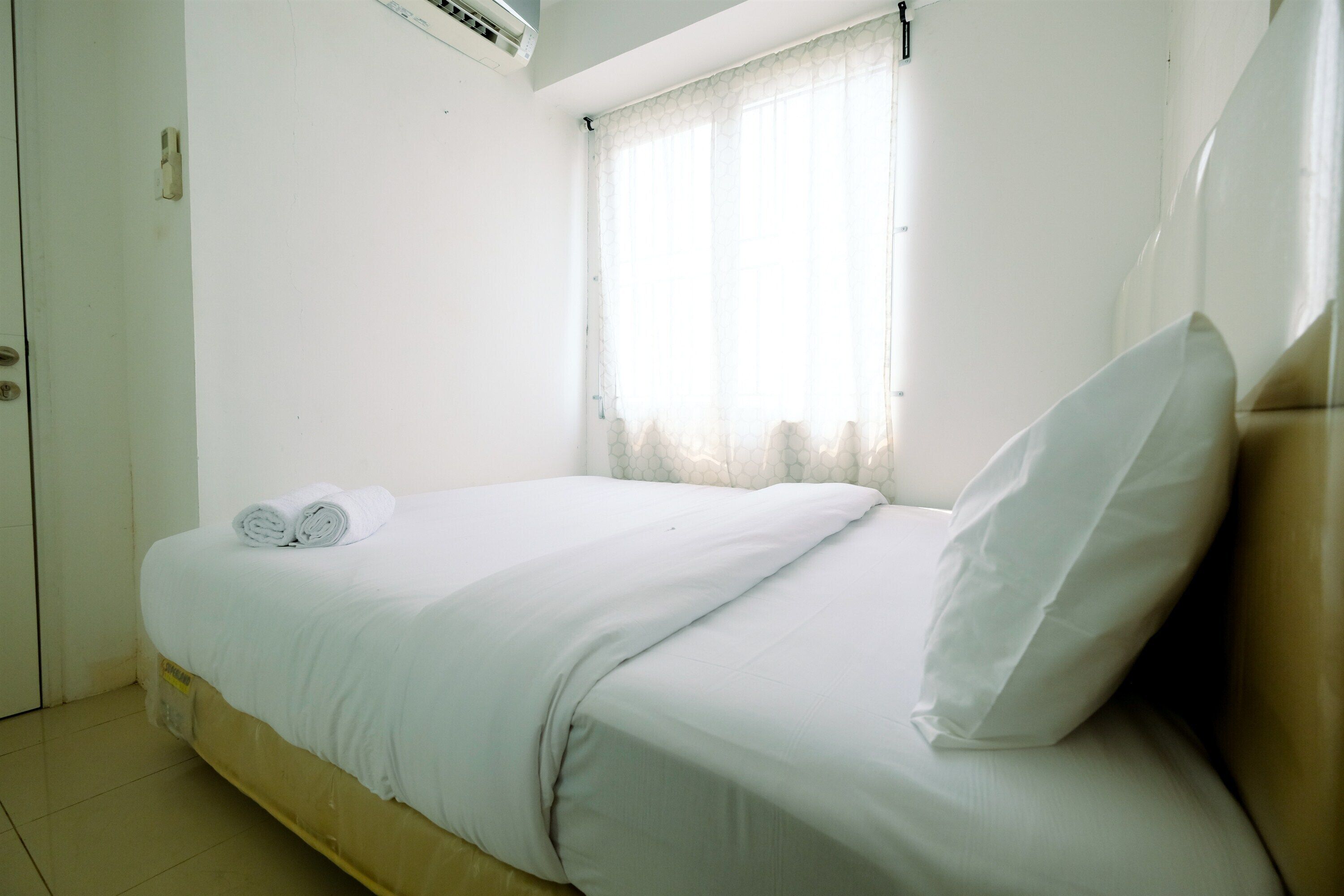 Una cama individual con sábanas blancas y una toalla enrollada, una ventana con cortinas translúcidas y una pared blanca.