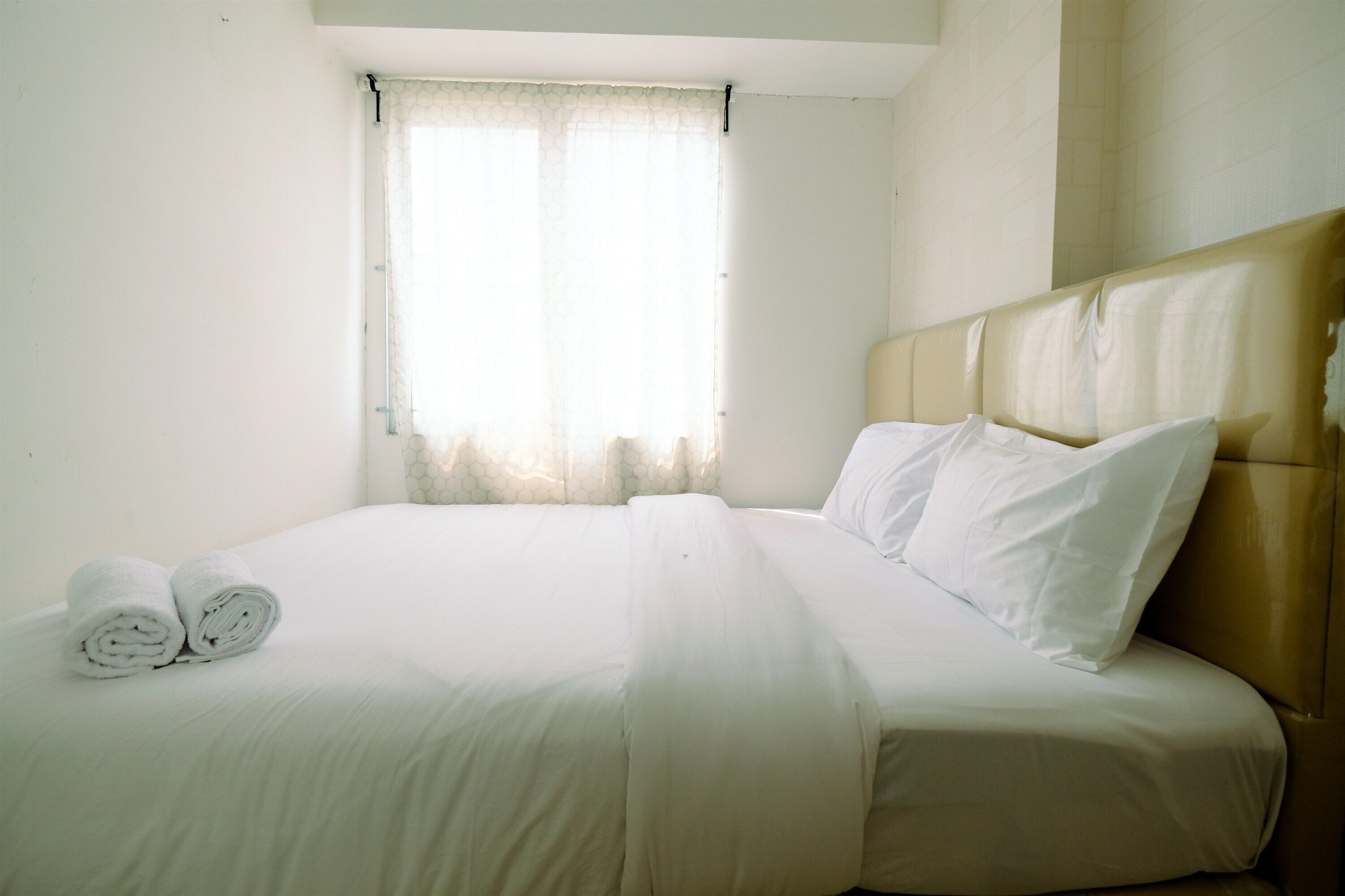 A neatly made bed with white linens and two rolled towels, a window with sheer curtains, and a headboard with a light-colored upholstered panel.