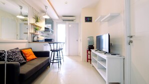 A modern living room with a sofa, TV, and a small kitchen area in the background. - Best Price 2BR Bassura City Apartment (Jakarta)