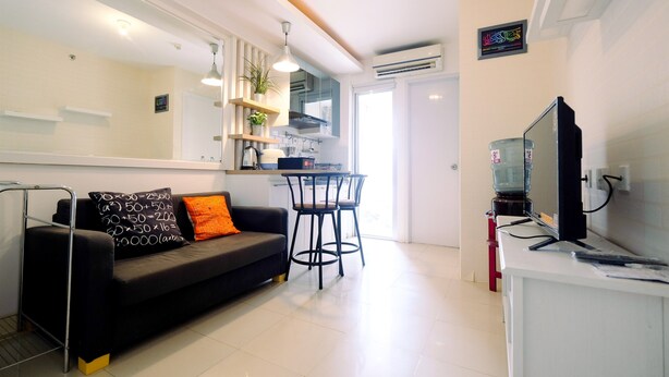 A modern living room with a black sofa, orange cushions, a dining area with two chairs, a TV on a white stand, and a kitchen area with a water dispenser. - Best Price 2BR Bassura City Apartment (Jakarta)
