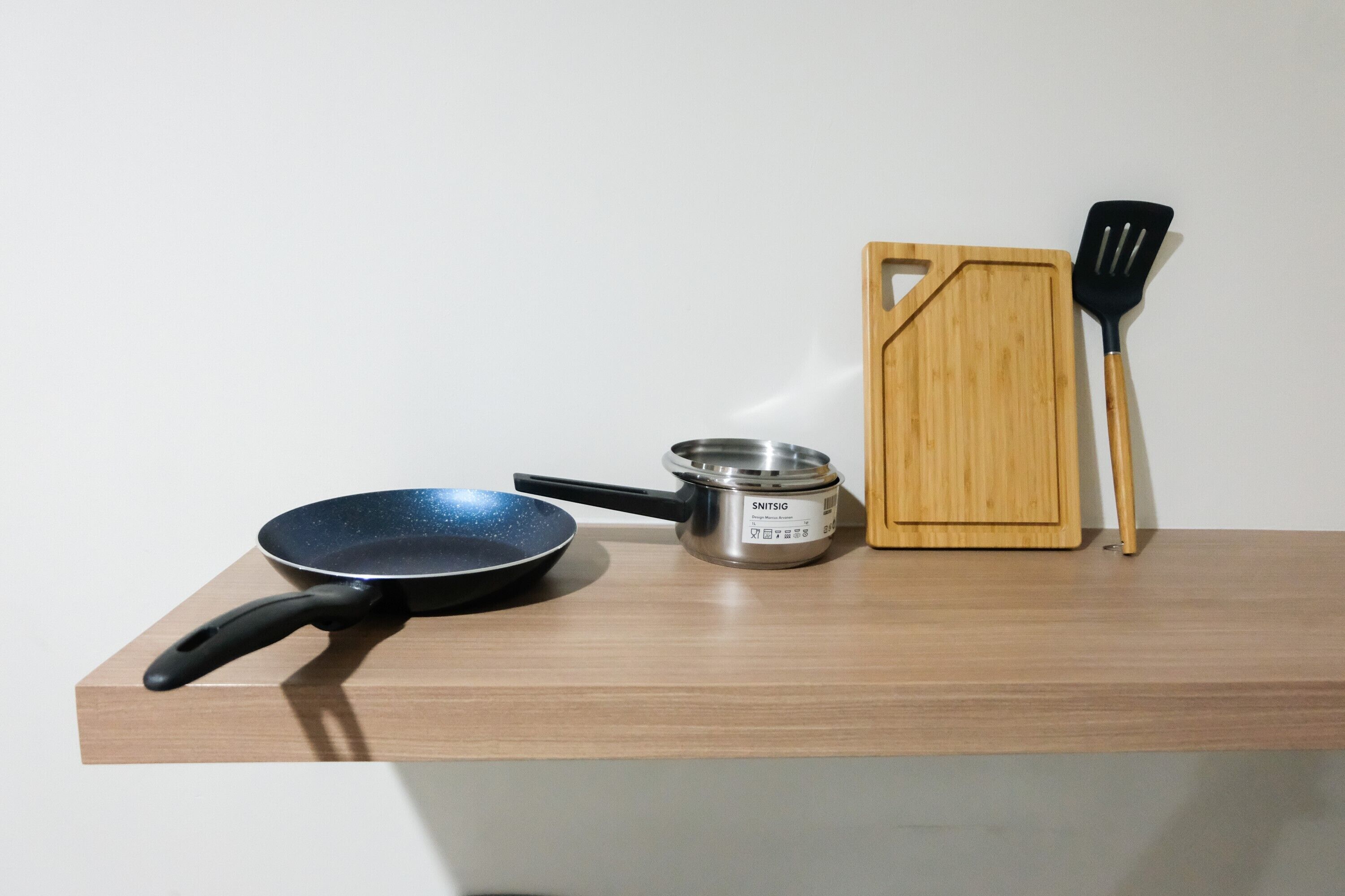 A kitchen counter with a frying pan, a pot, and a wooden cutting board with a spatula.