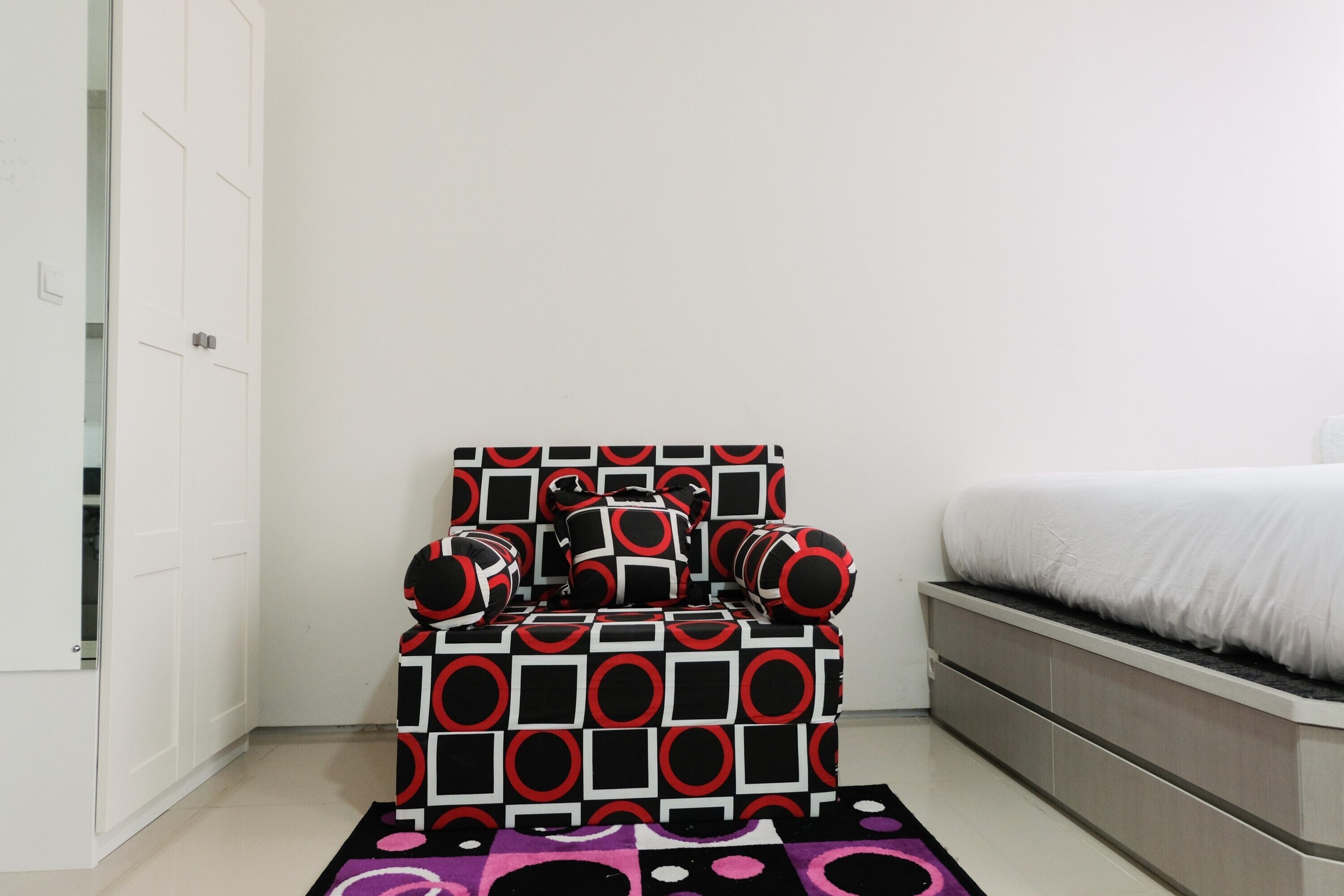 A bedroom with a bed, a chair with a geometric pattern, and a white wardrobe.