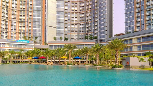 A large indoor swimming pool surrounded by palm trees and lounge chairs, with high-rise buildings in the background. - Wonderful 2BR at Gold Coast Apartment Pantai Indah Kapuk (Jakarta)