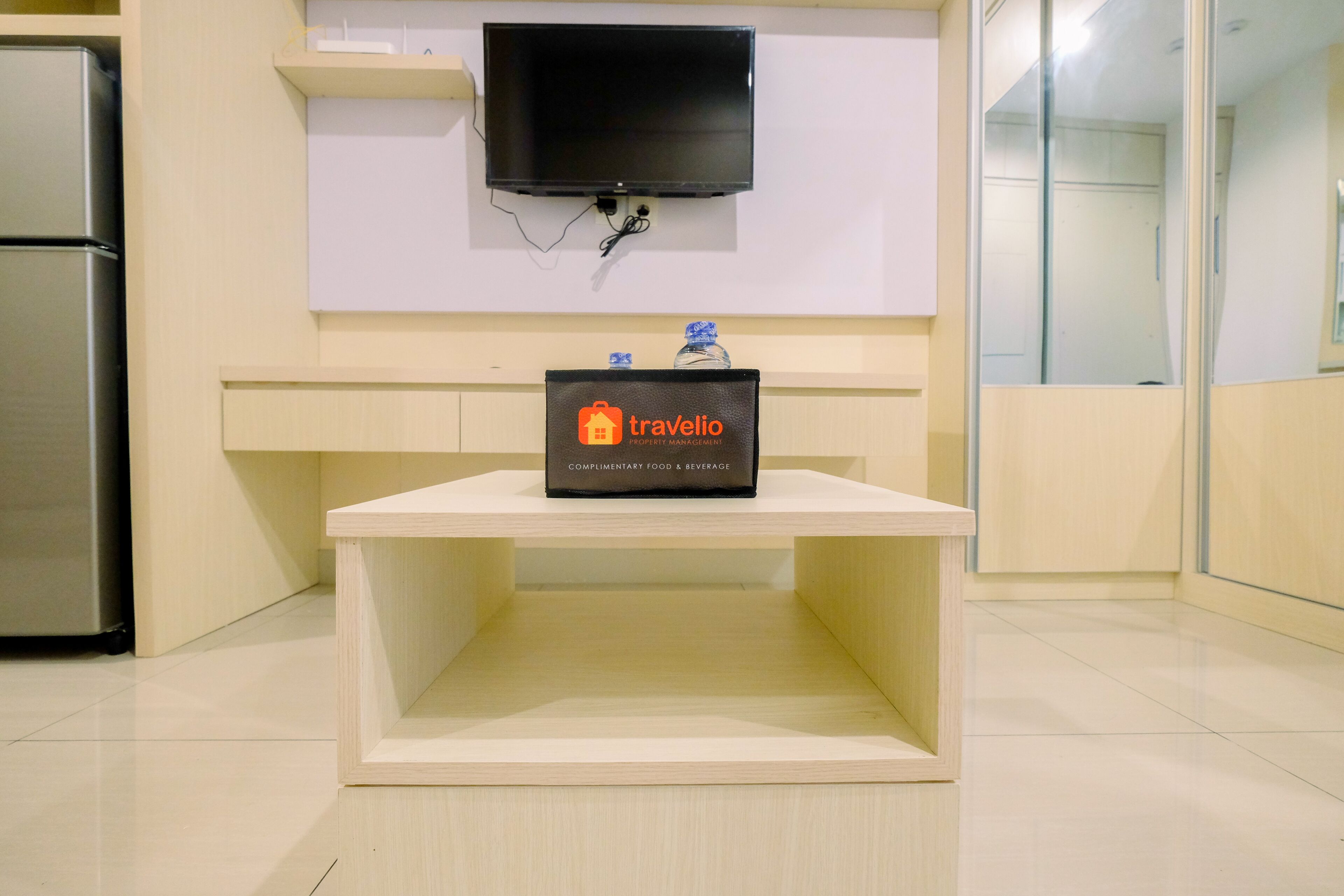 A traveler's desk with a flat-screen TV mounted above it, a complimentary food and beverage sign, and a refrigerator to the left.