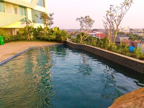 A swimming pool with tiled edges, surrounded by greenery and a building with large windows. - Modern 2BR Grand Kamala Lagoon Apartment (Bekasi)