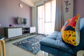 A living room with a sofa, a flat-screen TV, a floral rug, and a window with curtains. - Modern and Cozy 2BR Royal Olive Apartment (Jakarta)