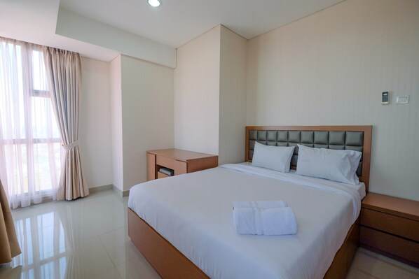 A hotel room with a large bed, a bedside table, a nightstand, and a window with curtains. - Modern and Cozy 2BR Royal Olive Apartment (Jakarta)