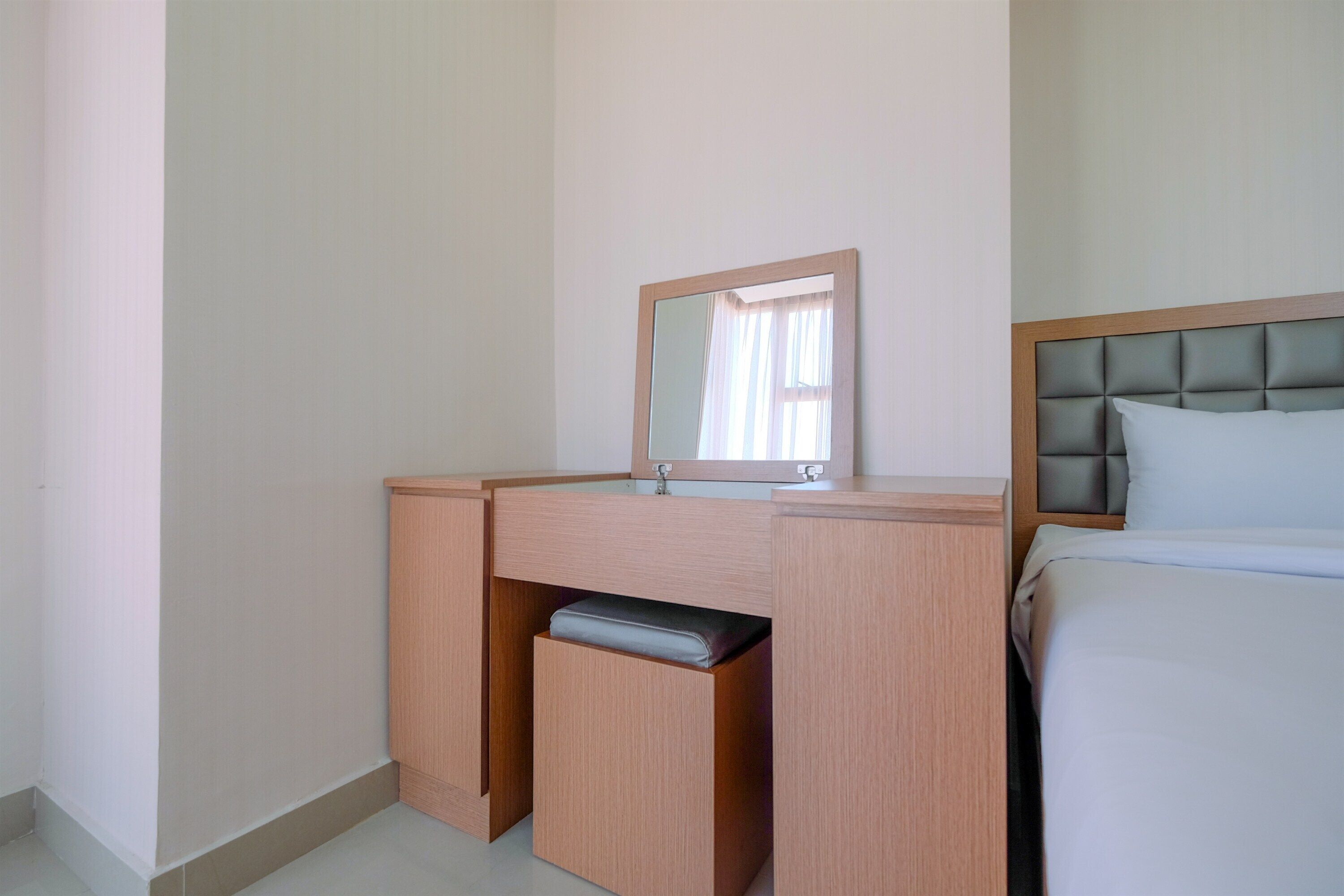 Een hotelkamer met een houten bureau, een spiegel, een bed met een grijze hoofdeinde en een lichtgekleurde muur.