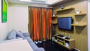 A modern living room with a flat-screen TV, grey sofa, and wooden shelves. - Modern 2BR at The Wave Epicentrum Apartment near Kuningan (Jakarta)