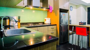 A compact kitchen with a sink, stove, and refrigerator. There is a clock on the wall and a red chair. - Modern 2BR at The Wave Epicentrum Apartment near Kuningan (Jakarta)