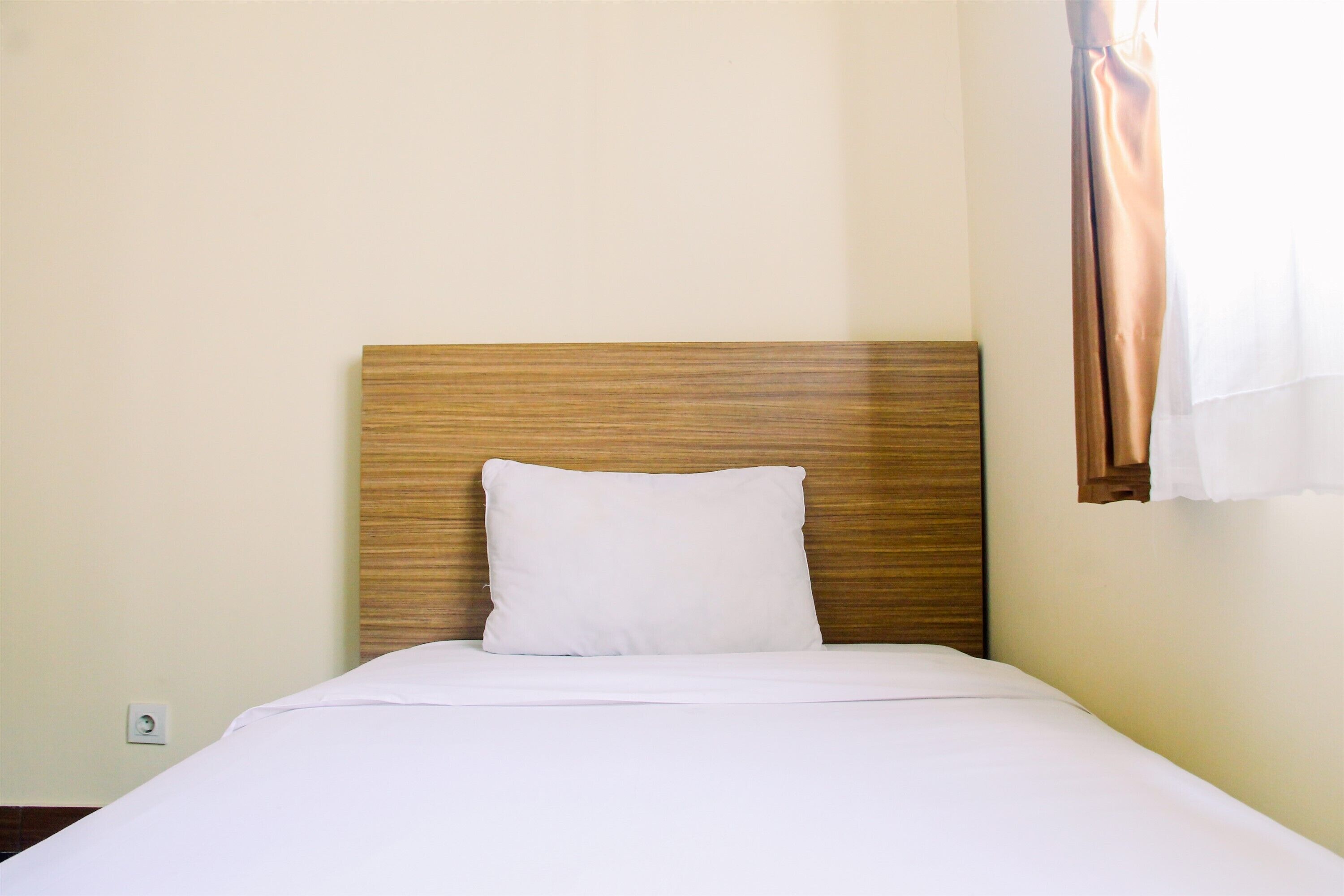 A neatly made bed with a wooden headboard, white bedding, and a power outlet on the wall.
