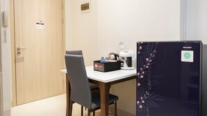 A hotel room with a wooden door, a white table with a chair, a coffee maker, a box labeled 'travello', and a refrigerator with a 'SHARP' logo. - 2BR Apartment at Elpis Residence near Ancol and Kemayoran (Jakarta)