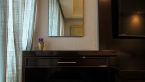 Habitación de hotel con un escritorio de madera oscura, un taburete pequeño, un florero con flores moradas, un espejo y un televisor montado en la pared.