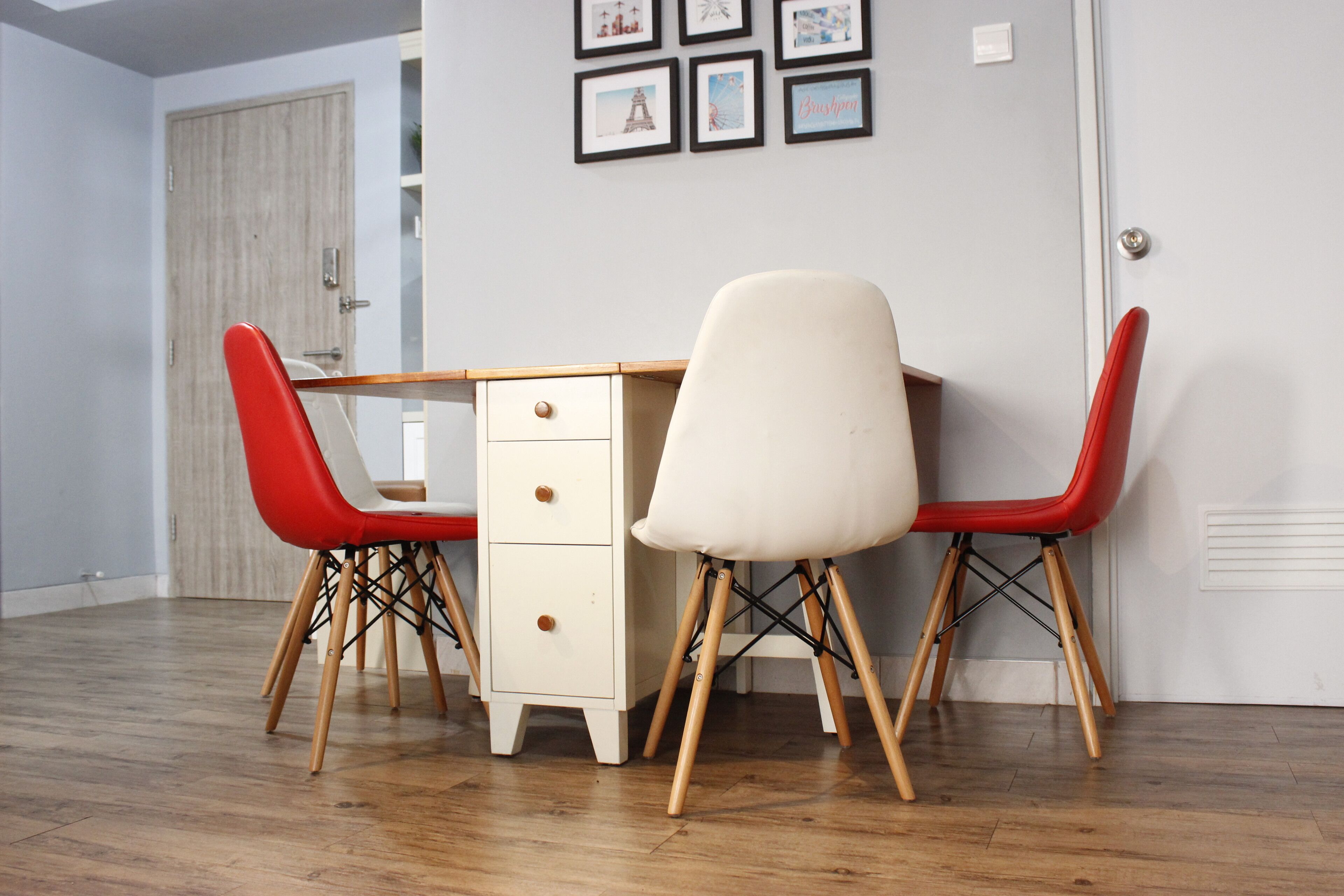 A dining area with a wooden table and two chairs, one red and one white, in a room with wooden flooring and a door in the background.