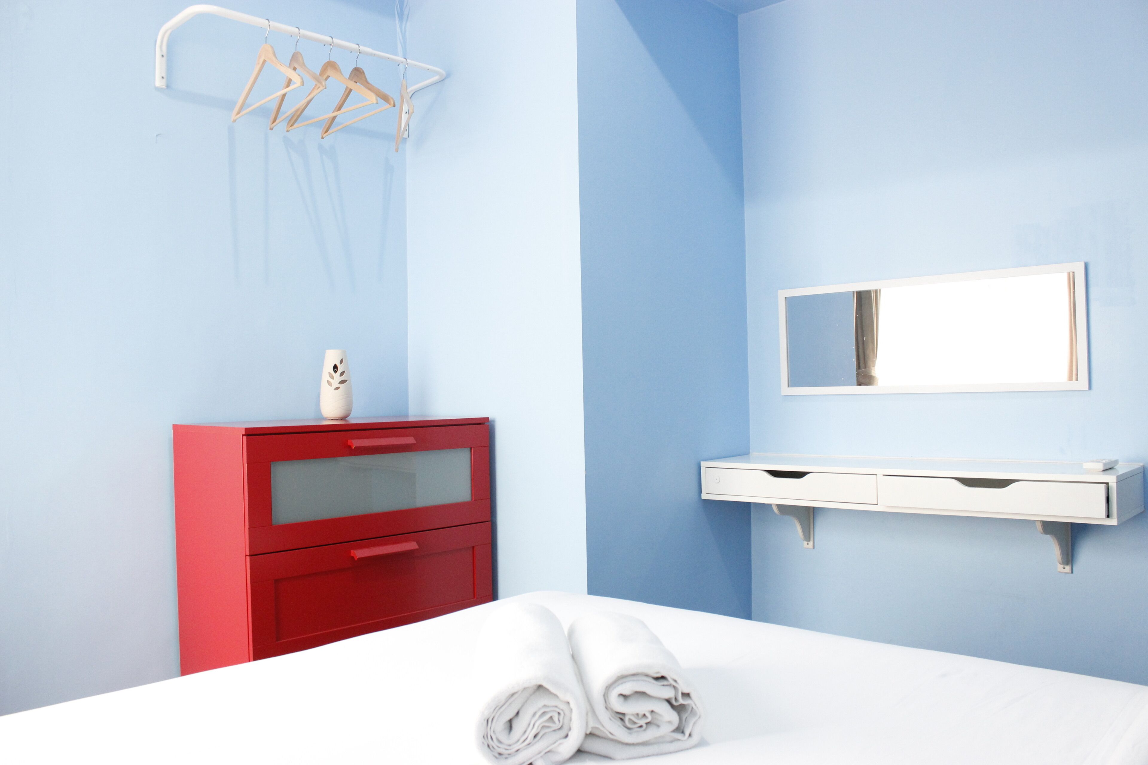 A hotel room with a bed, a red dresser, a white bedside table, a mirror, and a towel rack.