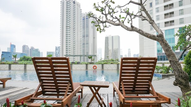 A poolside area with wooden lounge chairs and a table, surrounded by greenery and a view of tall buildings.