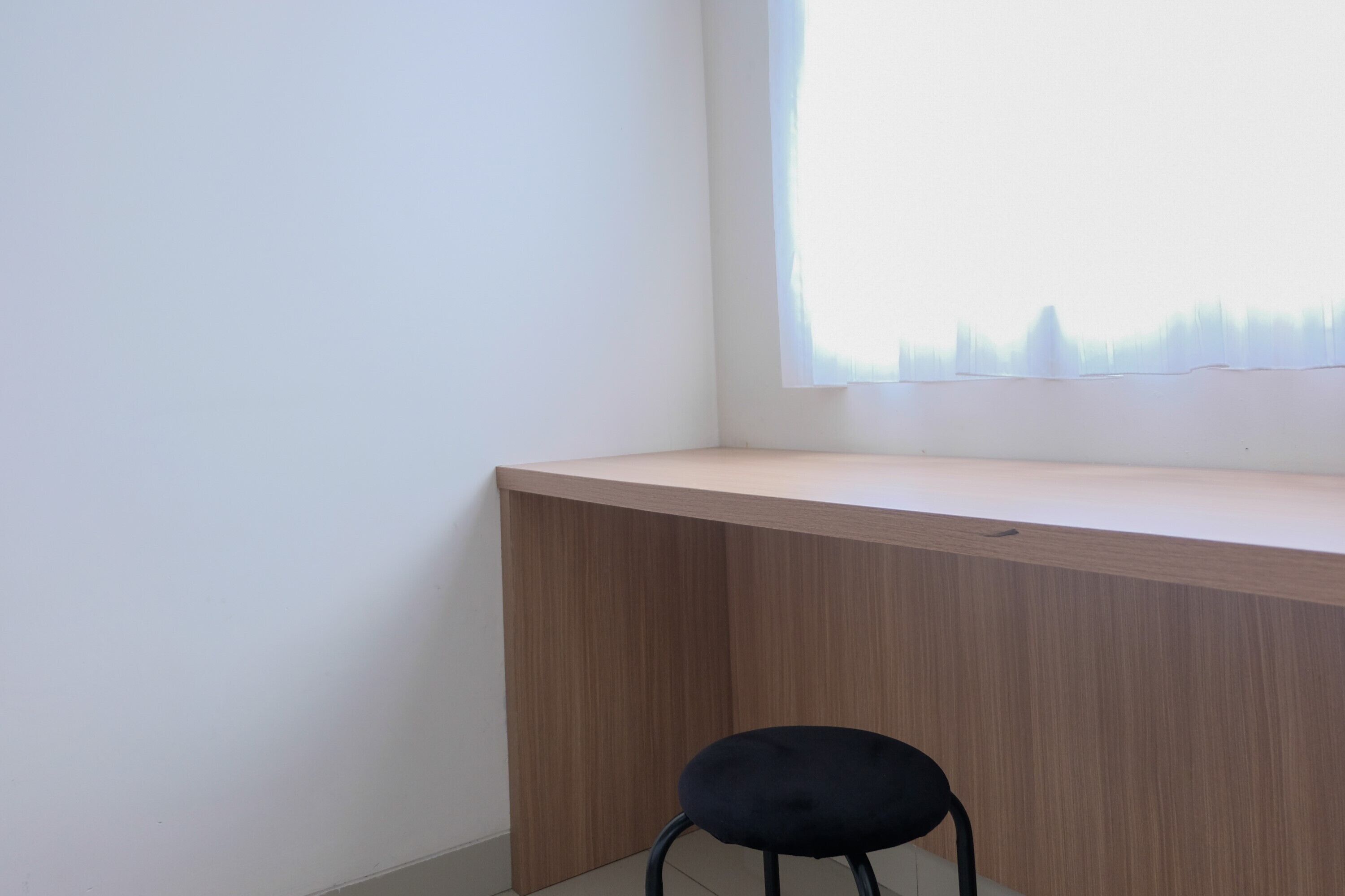 A corner desk with a chair, a window with sheer curtains, and a plain wall.