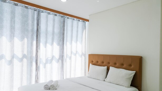 A neatly made bed with white linens and pillows, a wooden headboard, and a window with sheer curtains. - Modern and Cozy 1BR Brooklyn Alam Sutera Apartment (South Tangerang)