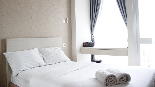 A neatly made bed with white linens, a bedside table with a lamp, and a window with curtains. - Classic Studio Room Taman Melati Apartment Jatinangor near UNPAD (Hegarmanah)