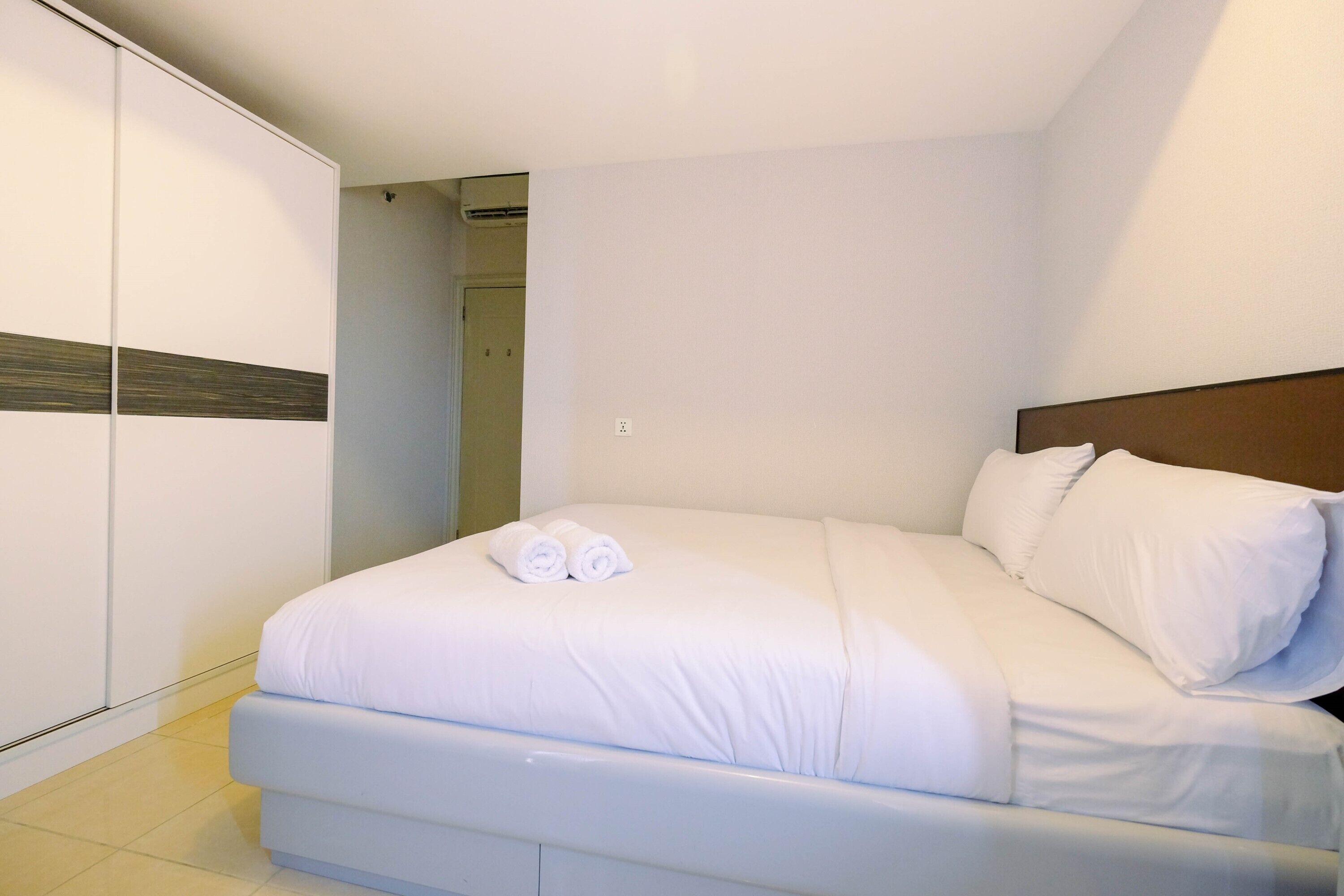 Un lit bien fait, avec du linge de lit blanc et deux serviettes enroulées, une tête de lit en bois et une armoire blanche à rayures horizontales.