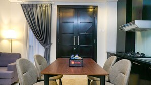 Quarto de hotel com uma mesa de jantar de madeira, quatro cadeiras, um sofá, um abajur, uma cozinha com fogão e uma porta preta.