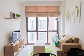 A modern living room with a sofa, TV, and a window with blinds. - Minimalist and Cozy Room 1BR Asatti Apartment (Pagedangan)