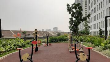 An outdoor exercise area with fitness equipment, surrounded by greenery and buildings in the background.