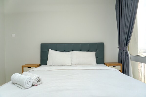 A neatly made bed with white linens and pillows, a dark green upholstered headboard, and a view of a cityscape through a window with grey curtains. - Cozy 2BR @ The Empyreal Condominium Epicentrum Apartment (Jakarta)