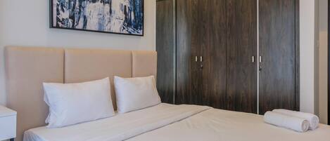 A neatly made bed with white linens, a large dark wooden wardrobe, and a framed abstract painting on the wall.