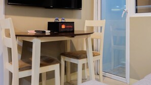 Een kamer met een wandgemonteerde flatscreen televisie, een kleine tafel met twee stoelen, een waterfles en een reistas.