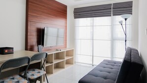 A modern living room with a sofa, a TV mounted on a wooden panel, a dining table with chairs, and a large window with blinds. - Elegant 1BR Apartment at Casa De Parco near AEON Mall (Pagedangan)