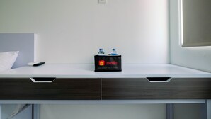 A hotel room with a white bed, a wooden desk with a black bag labeled 'Travelio' and two bottles, and a white wall with a power outlet. - Affordable Price Studio at Jababeka Riverview Apartment Cikarang (Cikarang)