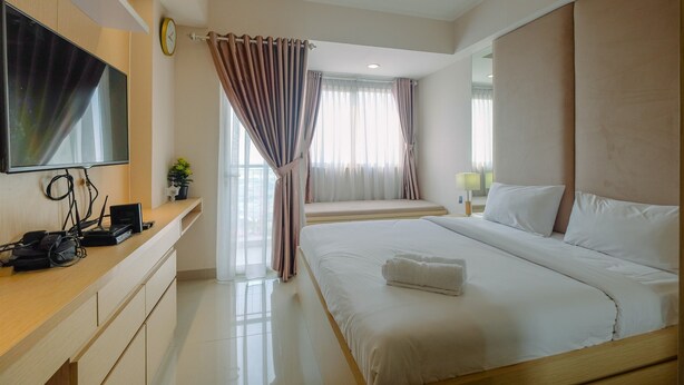 A modern hotel room with a large bed, a flat-screen TV mounted on the wall, a desk with a computer and phone, and a window with curtains. - Exclusive Studio at The Oasis Cikarang Apartment (Cikarang)