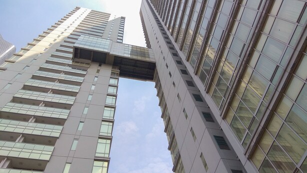 Un edificio alto y moderno con fachada de vidrio y una característica arquitectónica distintiva que recuerda a un puente o voladizo.