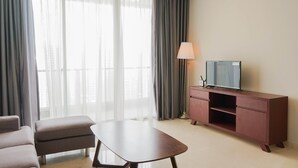 A modern hotel room with a sofa, a small table, a TV on a wooden cabinet, and large windows with sheer curtains. - Exclusive 2BR @ The Empyreal Condominium Epicentrum Apartment (Jakarta)