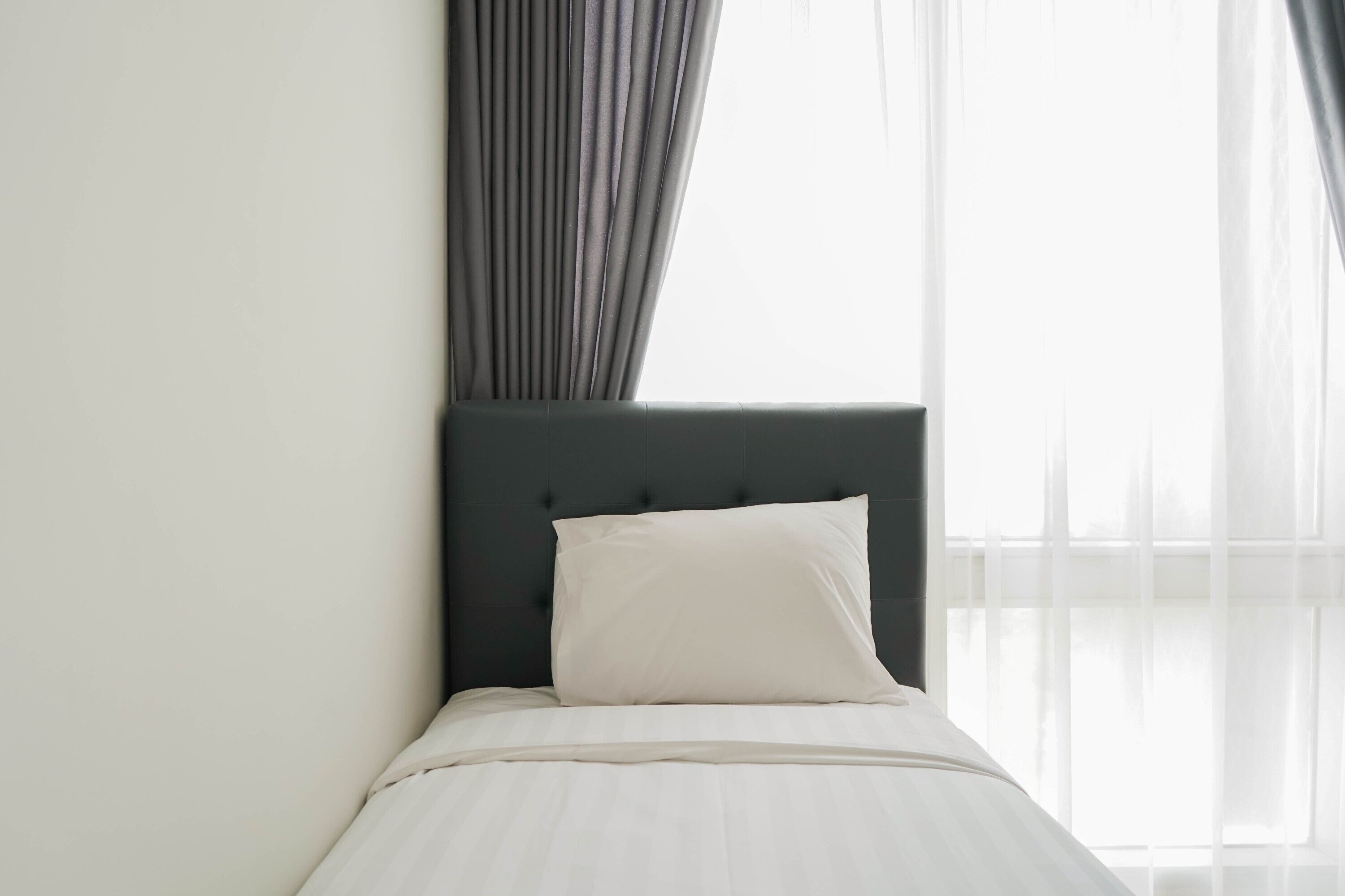 Cama bem arrumada com cabeceira escura e roupa de cama branca em um quarto com cortinas leves e uma janela grande.