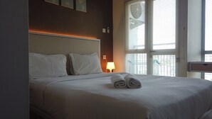 A neatly made bed with white linens, a folded towel, a bedside lamp, and a window with blinds. - Cozy Studio near UNPAD at Taman Melati Apartment Jatinangor (Hegarmanah)