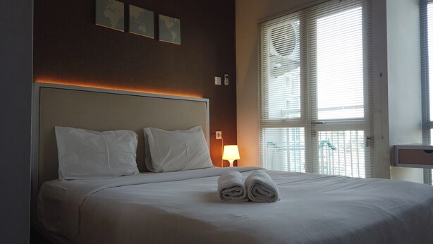 A neatly made bed with white linens, a bedside lamp, and a window with blinds. - Cozy Studio near UNPAD at Taman Melati Apartment Jatinangor (Hegarmanah)