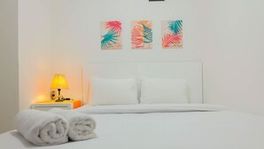 A neatly made bed with white linens and rolled towels, a bedside lamp, and three wall art pieces above the bed. - Pleasant 2BR Apartment at Bassura City near Shopping Mall (Jakarta)
