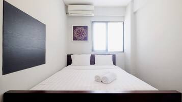 Habitación de hotel con cama, ropa de cama blanca, cabecera oscura, aire acondicionado montado en la pared, ventana con persianas y un adorno en la pared.