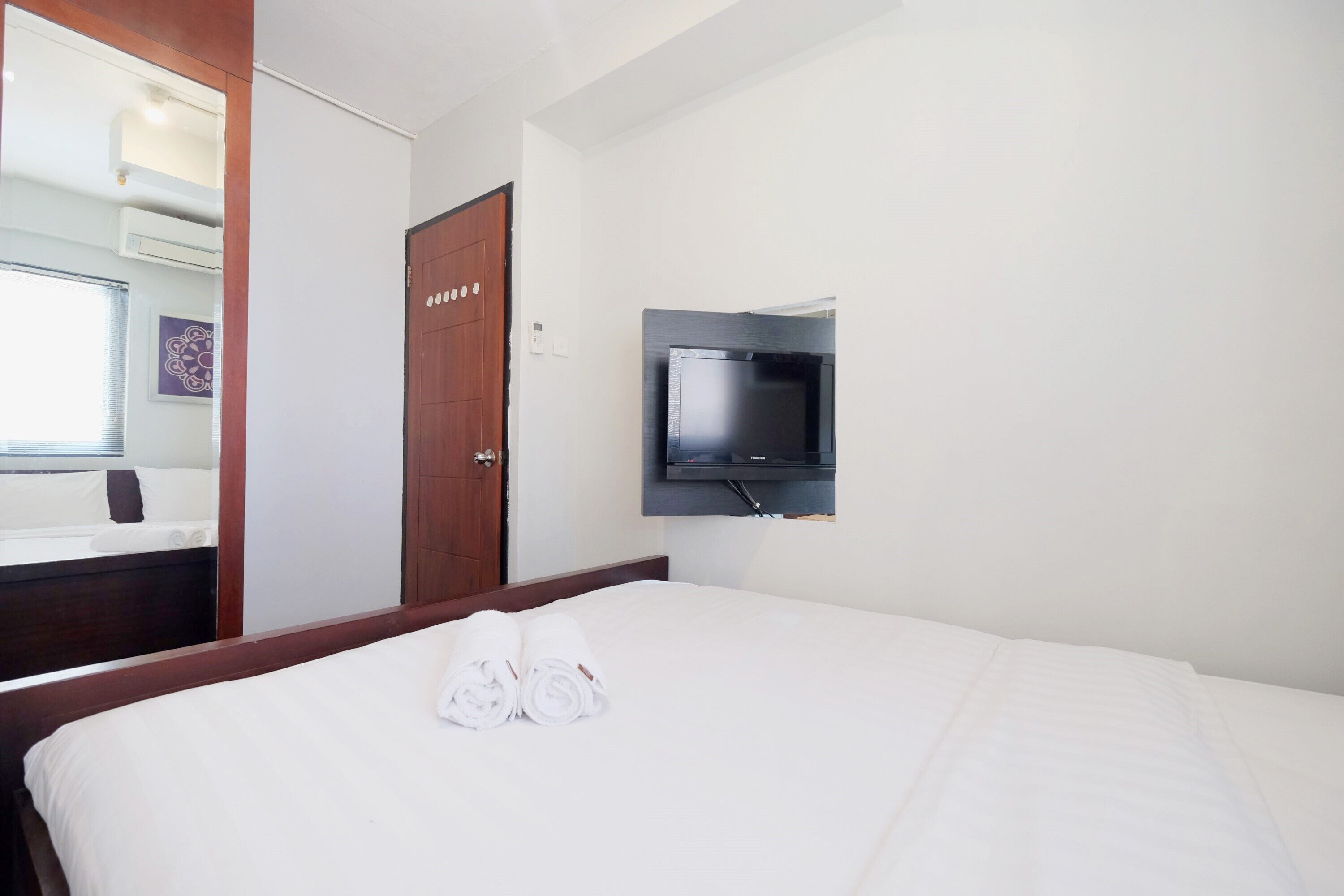 Habitación de hotel con una cama, un televisor montado en la pared, una puerta de madera y un espejo que refleja parte del cuarto.