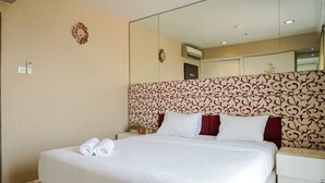 Una cama bien hecha con sábanas blancas y almohadas rojas, una mesita de noche con una lámpara y un reloj de pared.