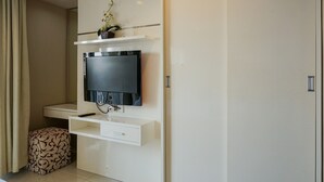 Una habitación moderna con un televisor montado, un pequeño estante con una planta, un escritorio blanco y un otomano estampado.