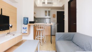 Una sala de estar moderna con un sofá gris, una mesita blanca pequeña y un televisor montado en la pared. Cuenta con una zona de cocina con un taburete de bar, un dispensador de agua y un salpicadero de azulejos.