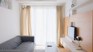 Una sala de estar moderna con un sofá gris, una mesa de centro blanca, un televisor montado y una ventana con cortinas traslúcidas.