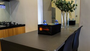 A kitchen area with a stove, a black box labeled 'Travelio' on the counter, and a vase with green leaves on the table. - Warm and Cozy 3BR Apartment at M-Town Residence (Gading Serpong)
