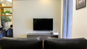 A living room with a flat-screen TV on a stand, a sofa, a dining table with chairs, and a bookshelf with plants. - Warm and Cozy 3BR Apartment at M-Town Residence (Gading Serpong)